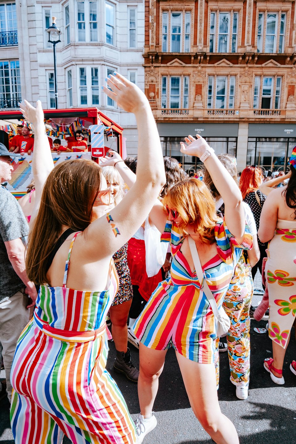 Two women in rainbow outfits dance at a street parade.