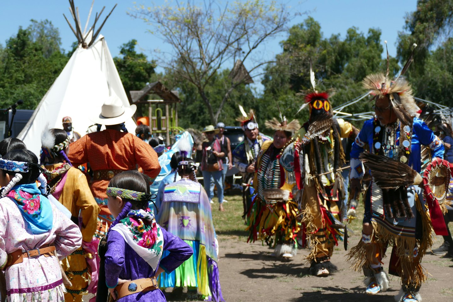 Indigenous people in traditional regalia gather outdoors near a teepee.